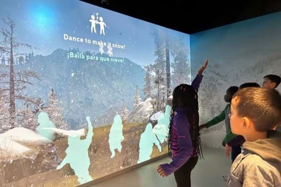 Children dancing in front of an interactive wall projection. The projection shows a mountain scene with falling snow, and silhouettes of the children appear to be in the scene.