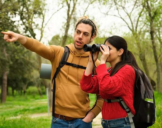 A man and woman look through binoculars while hiking through a forest