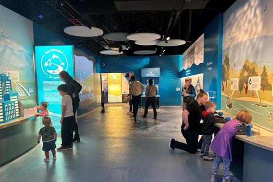 Families interact with exhibits inside a large room. The lighting in the room is low, drawing attention to the exhibits.
