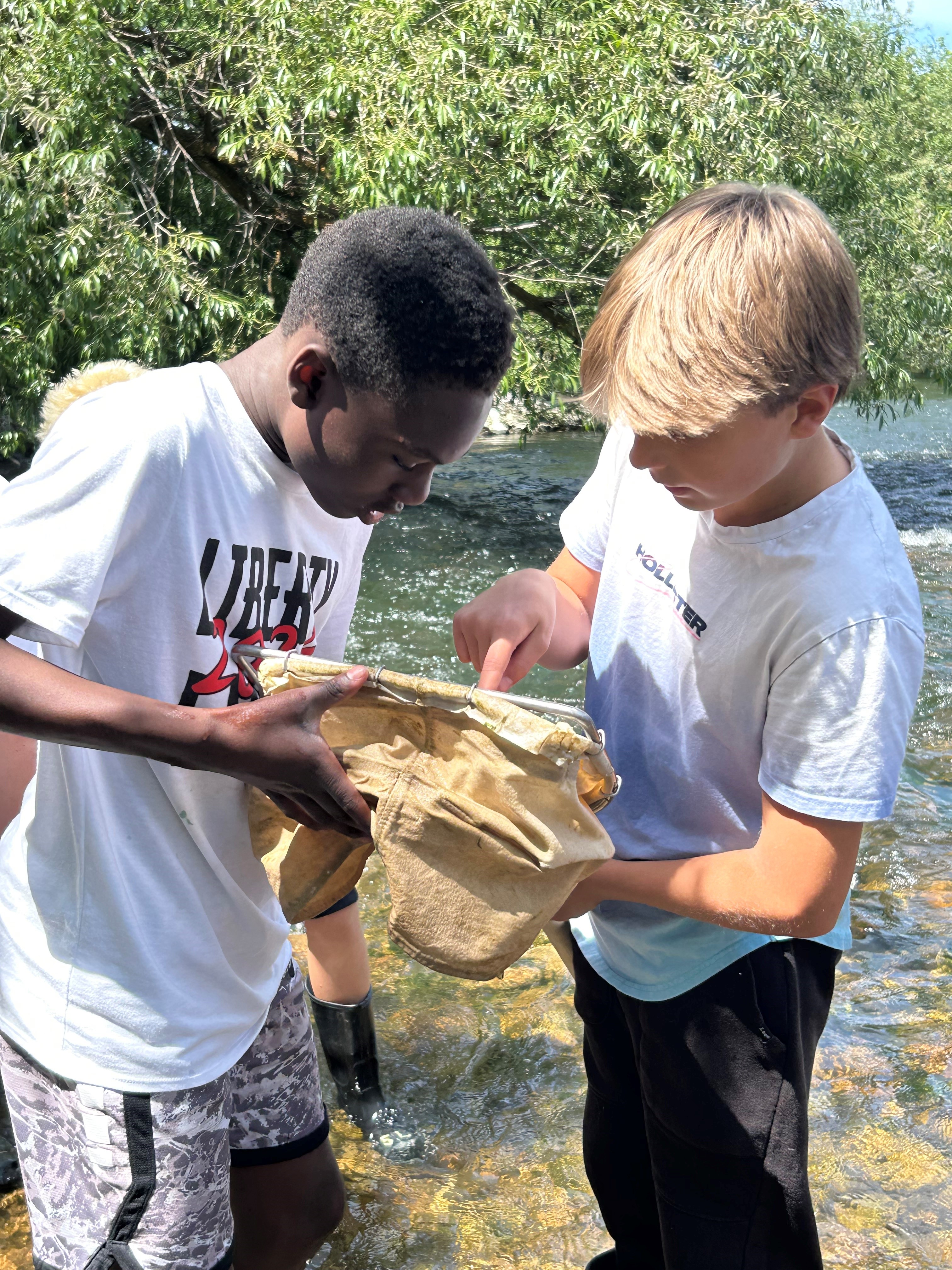 Two students stand in the river and examine specimens in a collection bag.