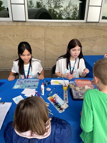 Two youth members of the Watershed Action Team educating young children and leading a craft at an event.