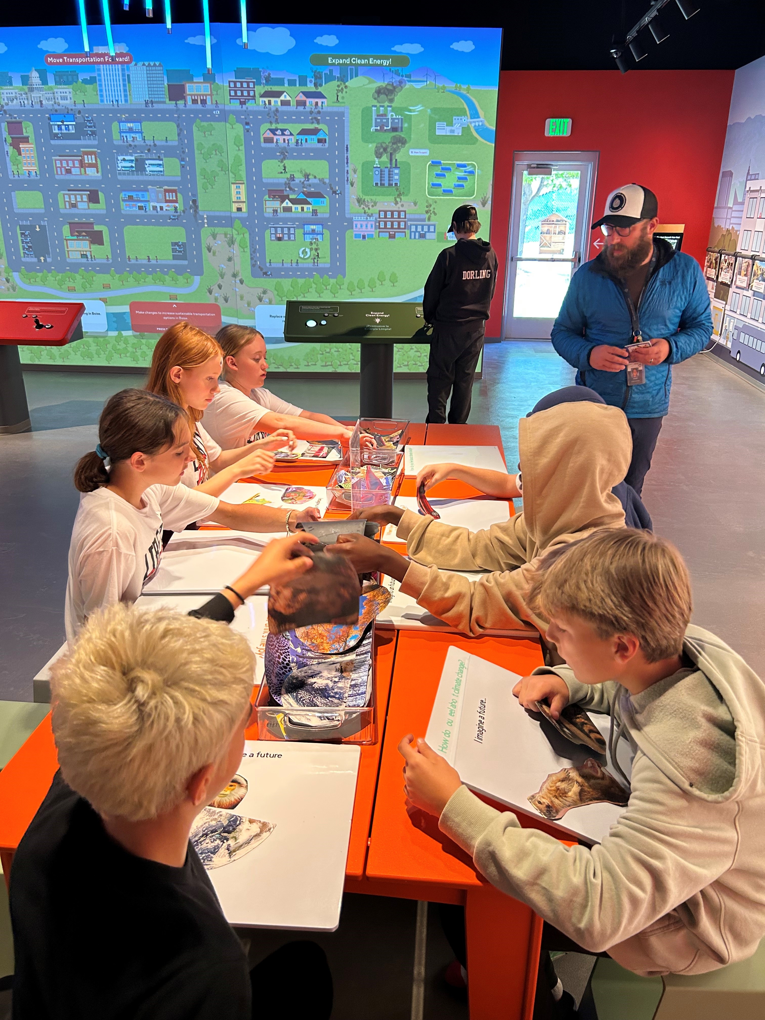 A group of students around a table using educational resources while an adult leader gives instructions.