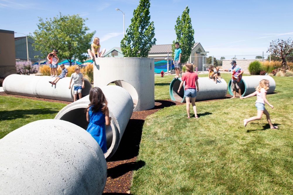 Concrete sewer pipes arranged into a playable area. The pipes laying form tunnels about three feet high. Several children are climbing through and on top of them.